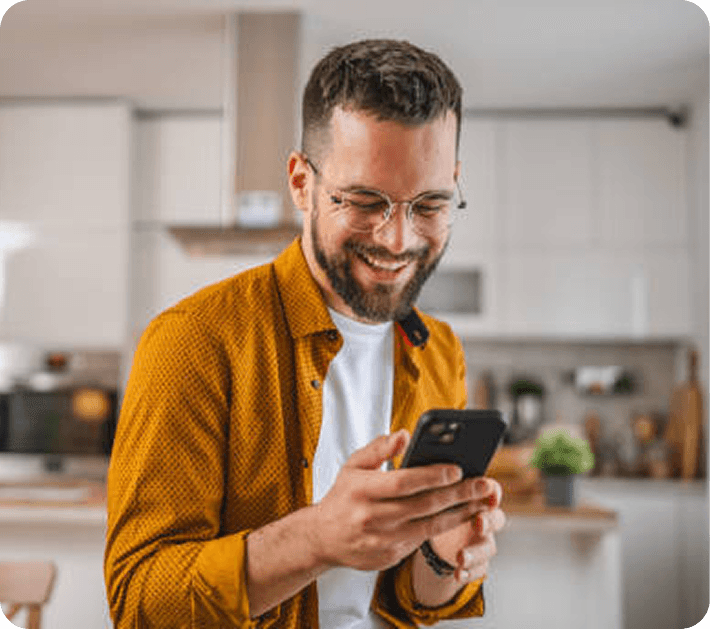 A man smiling at a text thread on his phone while standing in his kitchen